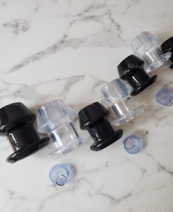 Angled close-up view of black and clear Hollow Anal Plugs in various sizes, featuring removable silicone stoppers, displayed on a marble surface.