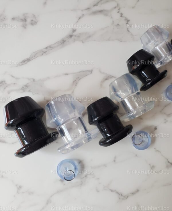 Close-up angled top view of black and clear hollow plugs in various sizes, featuring removable silicone stoppers, displayed on a marble surface.