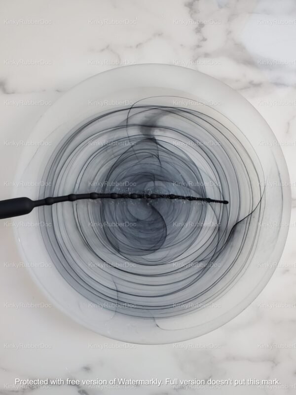 Black beaded silicone urethral vibrator submerged in circular sterilization bowl with swirling water patterns on marble surface
