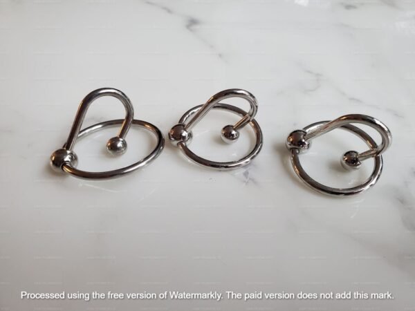 Three metal urethral plugs with ball tips and circular glans rings in different sizes on a marble surface