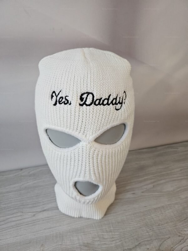 **Alt Text**: White knit balaclava with "Yes, Daddy!" embroidered in black text, featuring eye and mouth openings.