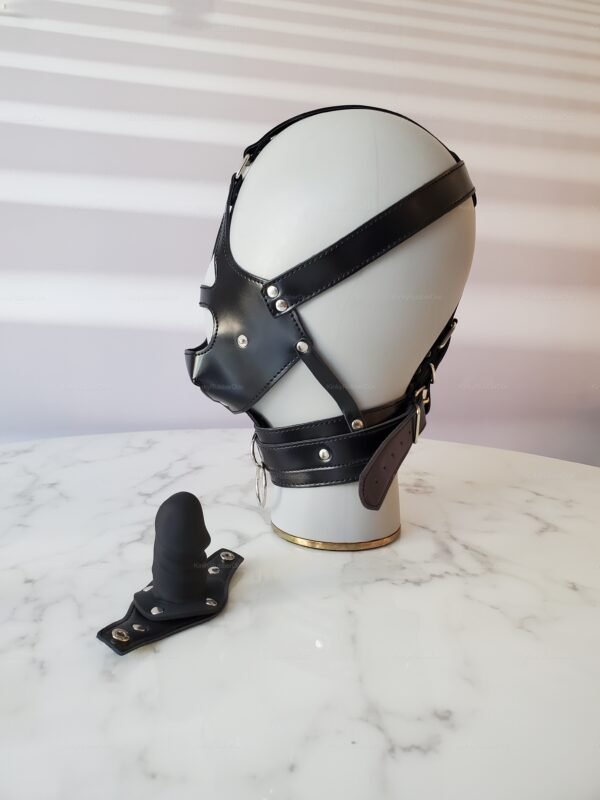 Black leather BDSM gag mask with straps and detachable mouthpiece on a marble surface.