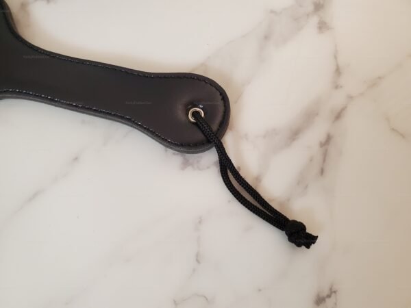 Black leather spanking paddle with paw design and wrist strap on a marble surface.