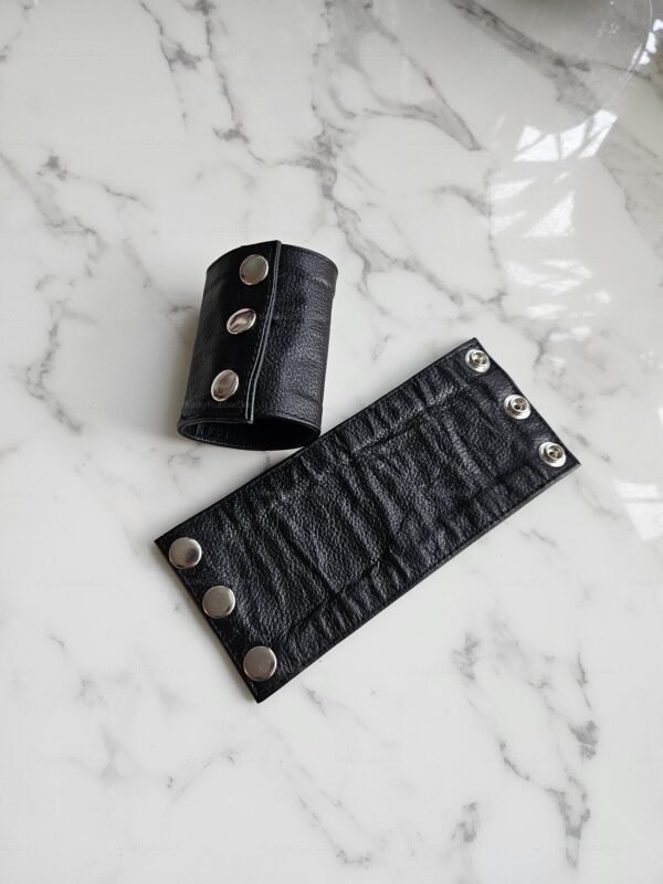 Black leather bracelet with snap closures, designed for a snug fit around the arm, lying on a marble surface.