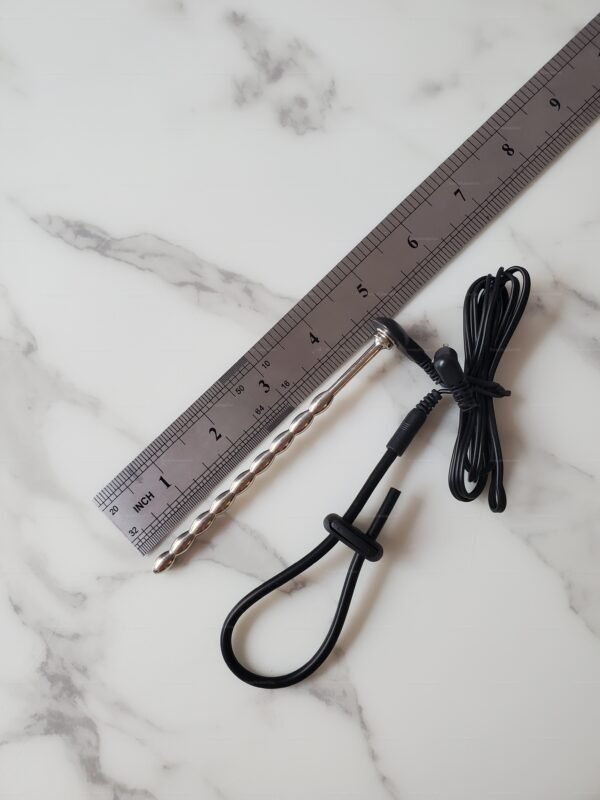 A sleek, metallic steel sounding penis plug with a textured design and an attached cable for e-stim play, placed next to a ruler on a marble surface.